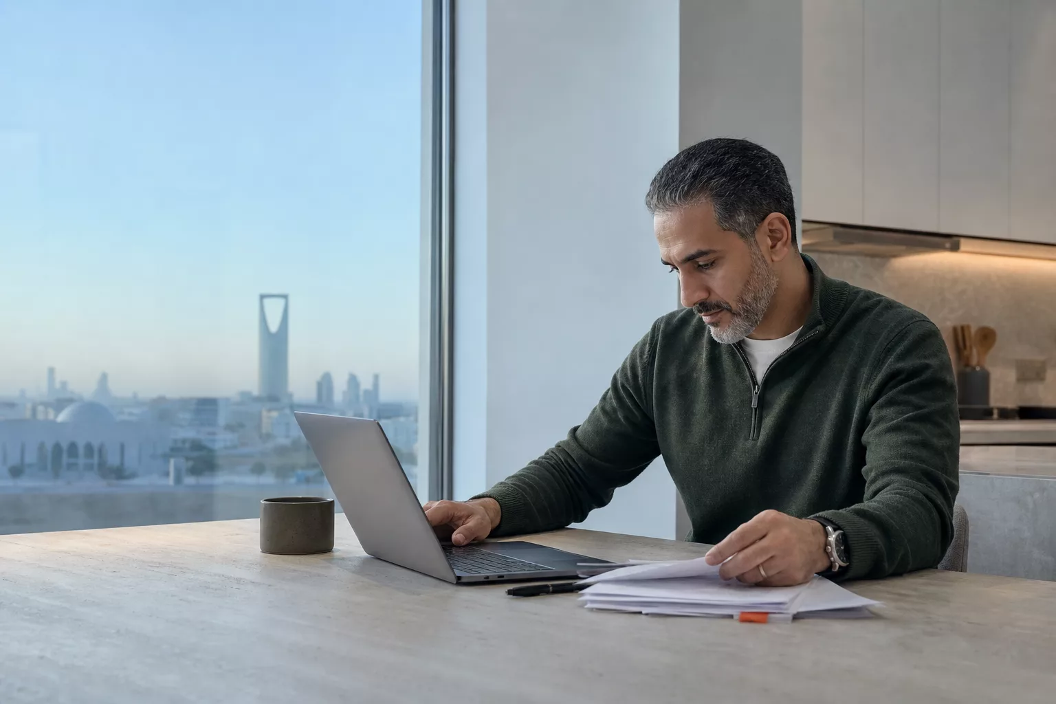 A senior professional at a light kitchen table with a laptop and printed pages in the quiet dawn.